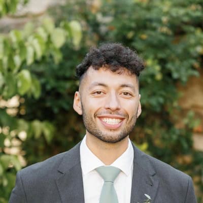 A person wearing a formal suit jacket, white shirt and light‑coloured tie is standing outdoors in front of leafy greenery.