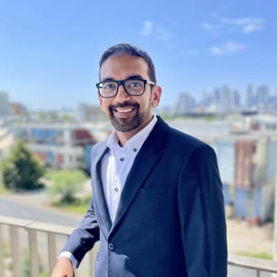 A person with dark hair and a beard with glasses wearing a dark blue suit and white shirt standing in front of some buildings with blue sky visible in the background.