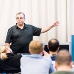 An individual stands at the front of a room giving a presentation to an audience. The individual gestures with one arm extended toward a screen or display. Several audience members are seated facing the presenter, and a blue sign with the word ‘European’ is visible.