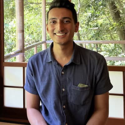 A person is seated indoors near large windows that look out onto greenery. The person is wearing a short‑sleeved button‑up shirt and is positioned in front of a wooden frame structure with natural light coming through.