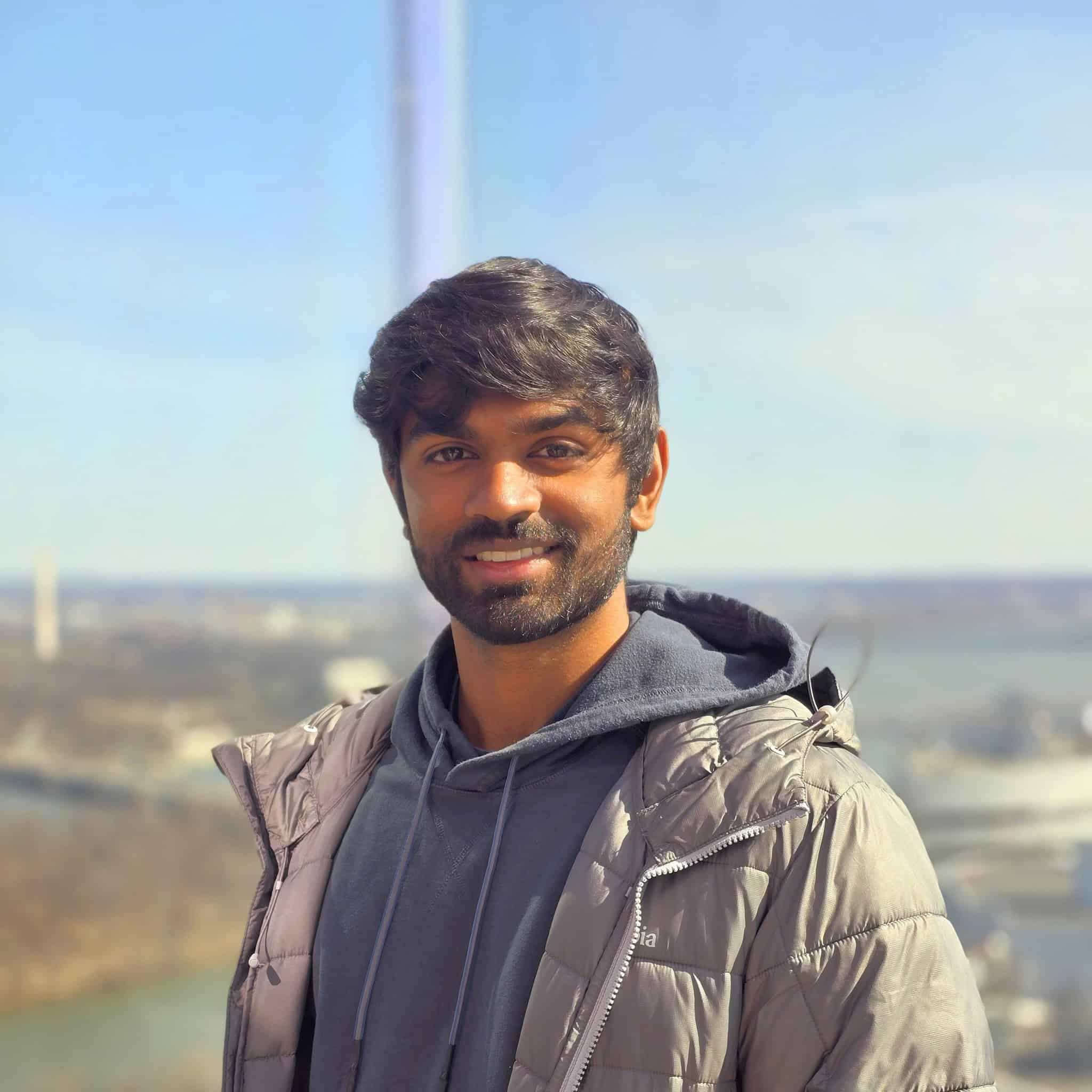 A person is outdoors wearing a light-colored padded jacket over layered clothing. The background features an open landscape with distant buildings, water, and a clear sky.