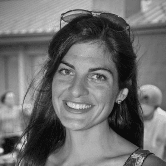 A person with long dark hair is shown outdoors in a black‑and‑white photograph. Sunglasses rest on top of the person’s head, and the background includes several other people and a building with a tiled roof.