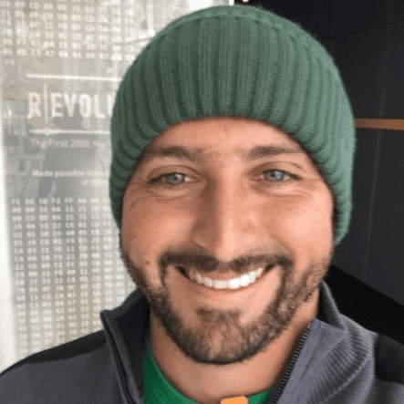 A smiling person with a dark beard wearing a green woolen hat.