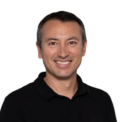 A smiling person with dark hair wearing a black polo shirt standing in front of a white background.