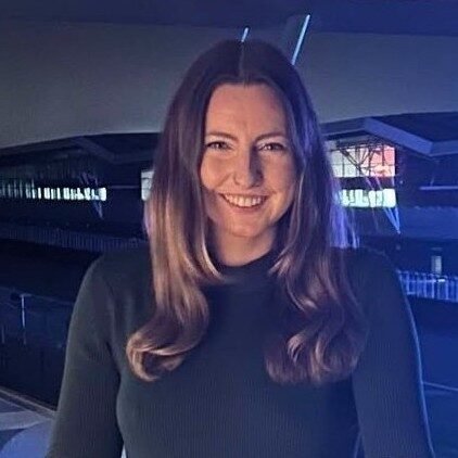 A person with long hair is shown from the waist up, wearing a dark long‑sleeved top and standing in a dimly lit environment with illuminated signs and architectural features in the background.
