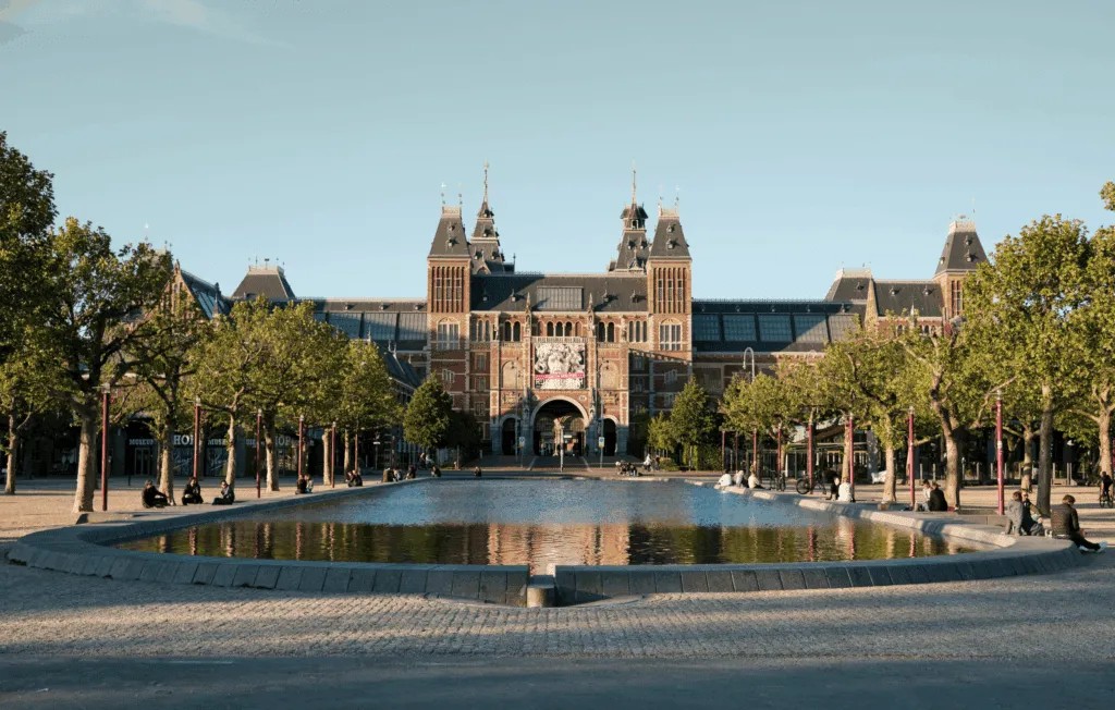 Front view of the Rijksmuseum with its large arched entrance, towers, and decorative façade, reflected in a long, shallow rectangular pool lined with trees