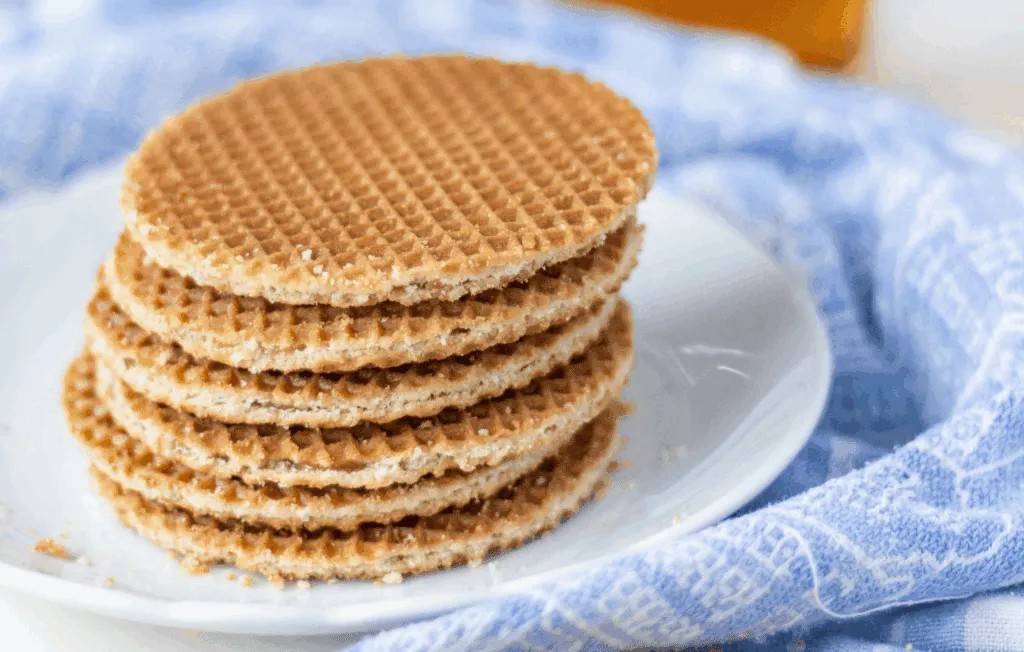Stack of round, thin stroopwafels with a caramel center placed on a white plate, set on a blue‑patterned cloth.