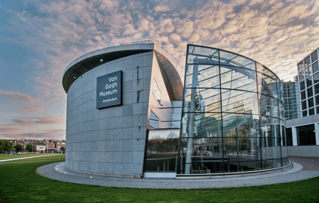 Exterior of the Van Gogh Museum featuring a curved grey building and an adjoining modern glass structure, set against a sky with scattered clouds.